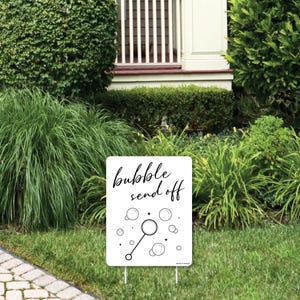May include: A white rectangular sign with the words "bubble send off" in black script, along with bubble graphics. The sign is on metal stakes, placed in a grassy yard with green bushes and a house in the background.