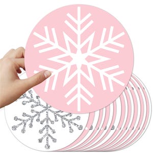 May include: A stack of pink and white snowflake-shaped decorations. One pink circle is held up, displaying a white snowflake design. A silver glitter snowflake is visible on a white circle. The decorations are suitable for winter or holiday crafts.