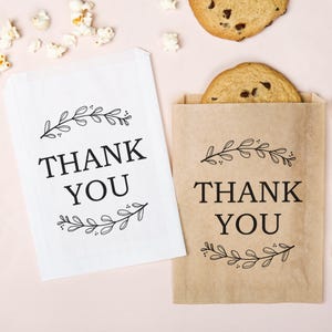 May include: Two paper treat bags, one white and one brown, with the words "THANK YOU" in bold black letters. Each bag has a decorative floral design above and below the text. A cookie is visible in the brown bag.