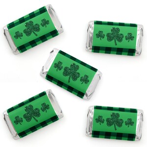 May include: Five green and black mini chocolate bar wrappers with a shamrock design. The shamrocks are green with glitter and the wrappers have a plaid pattern.