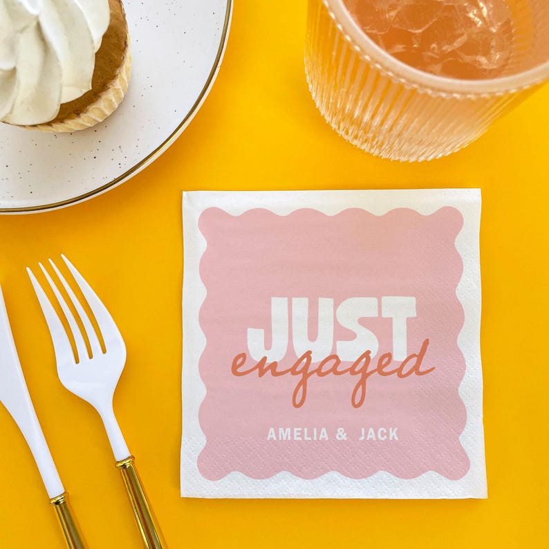 Op de afbeelding: Een witte vierkante servet met een roze geschulpte rand en de woorden "JUST engaged" in witte en koraalkleurige letters. De namen "AMELIA & JACK" staan eronder gedrukt. De servet ligt op een gele ondergrond, met een vork, mes en dessertbord.