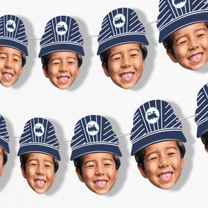May include: A string of paper cutouts featuring a smiling child's face wearing a navy blue train conductor hat with white stripes and a train logo. The hats are strung together on a white string, creating a festive banner.