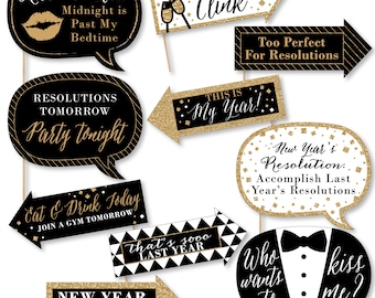 May include: A collection of black and gold photo booth props with various phrases for a New Year's Eve celebration. The props include speech bubbles and arrows with text like "Kiss Me Now," "Party Tonight," and "New Year, Same Me."