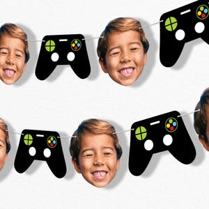 May include: A garland featuring alternating images of a smiling child's face and black video game controllers. The controllers have green, yellow, red, and blue buttons, with white and black accents. The garland is strung on a white string.