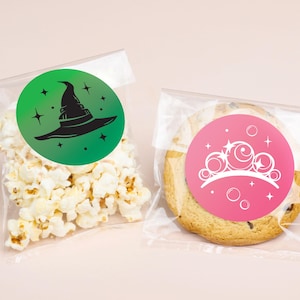 May include: Two clear plastic bags, one filled with popcorn and the other with a cookie. The popcorn bag has a green circular sticker with a black witch hat and stars. The cookie bag has a pink sticker with a white tiara and stars.