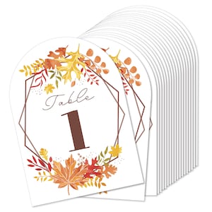 May include: Set of 12 white table number cards with a geometric frame and fall foliage design. The cards are shaped like an arch and have the text "Table" and a number. The number on the card shown is 1.