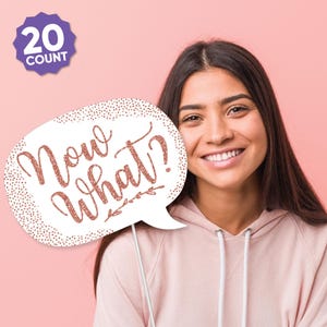 May include: A photo booth prop in the shape of a speech bubble with the words "Now What?" in rose gold glitter. The prop has a white background with a dotted border and is held by a person wearing a pink hoodie. A purple badge in the upper left corner says "20 Count".