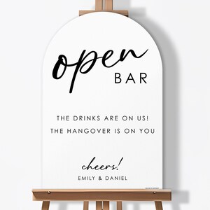 May include: A white sign with an arched top, displaying the words "open BAR" in black script. Below, it reads "THE DRINKS ARE ON US! THE HANGOVER IS ON YOU" and "cheers!" with names "EMILY & DANIEL". The sign is mounted on a wooden easel.