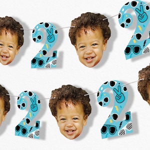 May include: A festive banner featuring the number "2" in light blue, adorned with sunglasses, lightning bolts, and peace signs. The banner also includes smiling faces of a young child, creating a cheerful and celebratory atmosphere.
