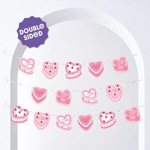 May include: A pink heart-shaped banner with the words "Happy Birthday" on some of the hearts. The banner is double-sided and has a cake design with white frosting and red cherries. The banner is strung on a white string.