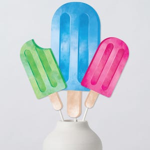 May include: Three colorful popsicle decorations in a white vase. The popsicles are green, blue, and pink, with wooden sticks. The green popsicle has a bite taken out of it. The popsicles have a watercolor effect.