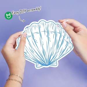 May include: A white and blue seashell-shaped craft with the text "Easy DIY assembly!" on a purple background. The craft is held by two hands, with a string attached to the top.