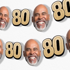 May include: A festive banner featuring the number "80" in gold glitter with a black outline, interspersed with smiling faces of a man with a gray beard. The banner is strung on a white string, perfect for an 80th birthday celebration.