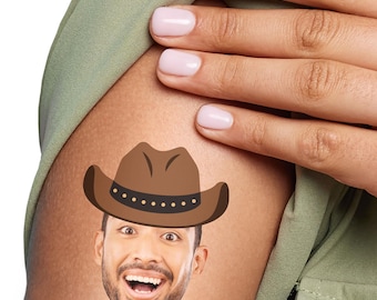 May include: A temporary tattoo of a smiling man's face wearing a brown cowboy hat with a black band and gold studs. The tattoo is on a person's arm, with a green shirt and a hand with pink nail polish in the background.
