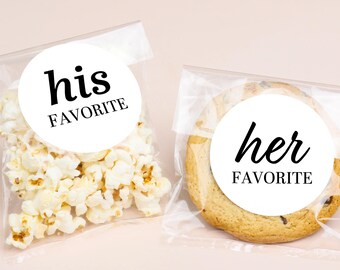 May include: Two clear cellophane bags, one filled with popcorn and the other with a large cookie. Each bag has a white circular sticker with black text. One says "his FAVORITE" and the other says "her FAVORITE".