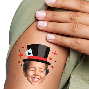 May include: A temporary tattoo featuring a cartoon illustration of a child's face wearing a black top hat with a red band and a playing card with a spade symbol. The tattoo is surrounded by red stars.