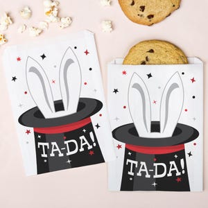 May include: Two white paper treat bags with a black top hat design, white rabbit ears, and the text "TA-DA!". The bags are decorated with red and black stars and dots. One bag contains a large cookie.
