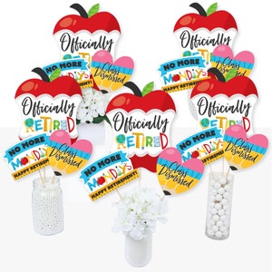 May include: A set of six red apple-shaped table centerpieces with the text "Officially Retired" and other retirement-themed phrases. The centerpieces are decorated with colorful pencils, a globe, and a ruler.