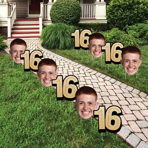 May include: A set of lawn signs featuring a repeating photographic cutout of a smiling person's face paired with a large, gold-colored number 16. Each sign is mounted on a thin metal stake, designed for placement in a grassy outdoor area.