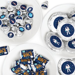 May include: A set of party favors featuring astronaut and space-themed stickers on chocolate candies. The candies are wrapped in silver foil and the stickers feature a blue background with stars, planets, and an astronaut. The stickers also include the text "Out of this World".