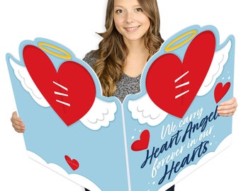 Puede incluir: Un gran recorte de cartón de un cielo azul con dos corazones rojos con alas y halos. Los corazones están cosidos juntos. El texto "We carry Heart Angel forever in our Hearts" está escrito en azul en el recorte.
