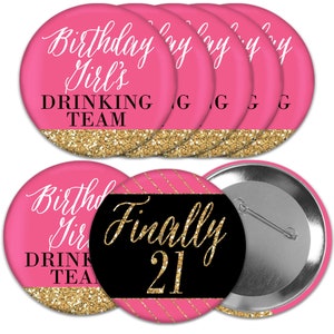 May include: A collection of pink and gold party buttons. The buttons feature the text "Birthday Girl's DRINKING TEAM" and "Finally 21" in white and gold script. The bottom of the buttons have a gold glitter design.