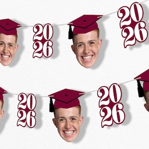 May include: A graduation banner featuring smiling faces wearing maroon graduation caps with black tassels. The banner also includes cutouts of the numbers "20" and "26" in a white font, celebrating the class of 2026.