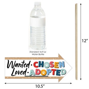 May include: A brown arrow-shaped sign with the text "Wanted. Chosen. Loved. Adopted." in colorful letters with paw prints. The sign is 10.5 inches wide and is propped up with a wooden stick that is 12 inches tall. A standard 16.9 oz water bottle is shown for size comparison.