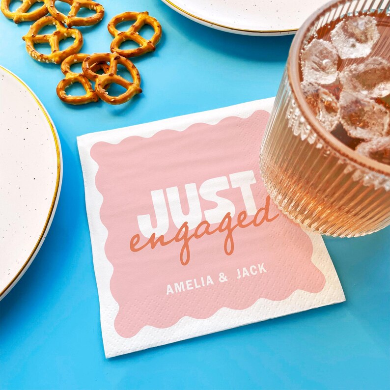 Op de afbeelding: Een roze en witte papieren servet met de woorden "JUST engaged" en de namen "AMELIA & JACK" erop gedrukt. Het servet ligt op een blauw oppervlak, met pretzels, een drankje en een bord zichtbaar.