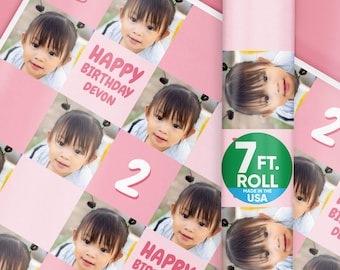 May include: Pink birthday wrapping paper featuring a repeating pattern of a young girl's photo, the number 2, and the text "HAPPY BIRTHDAY DEVON." A green sticker on the roll reads "7 FT. ROLL MADE IN THE USA."