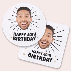 May include: Two white coasters with a smiling man's face and black burst lines. The text "HAPPY 40TH BIRTHDAY" is printed in black. One coaster is round, the other is square.
