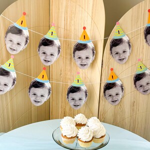 May include: A festive birthday banner featuring smiling baby faces wearing party hats with the number "1". The banner is strung across a wooden backdrop. Cupcakes with white frosting are on a glass plate.