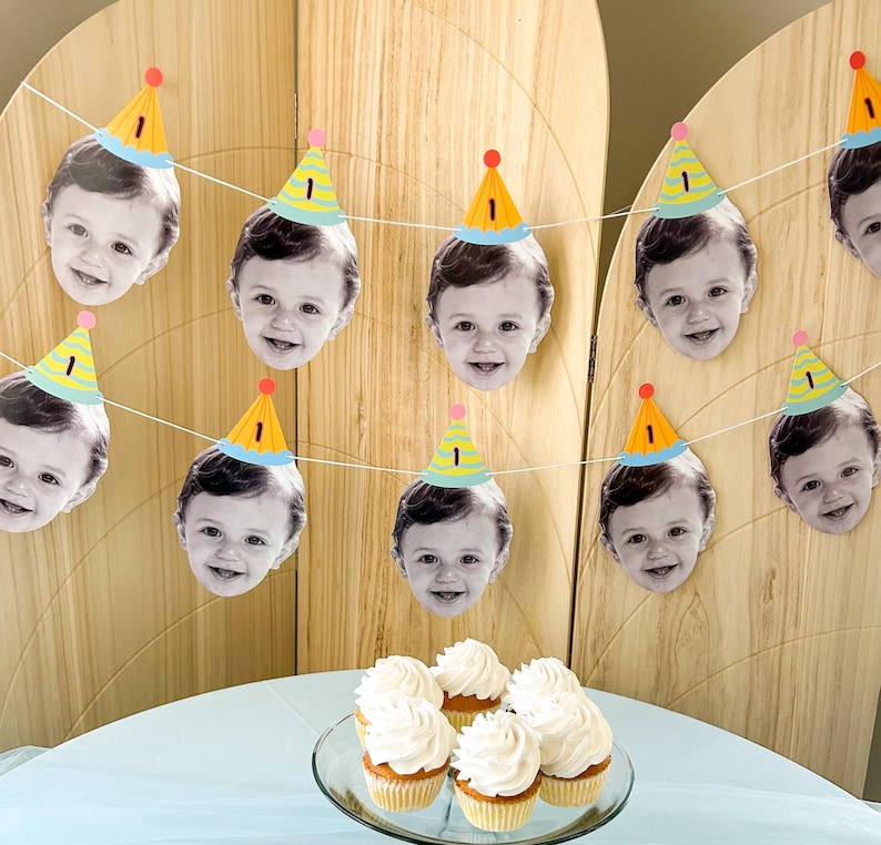May include: A festive birthday banner featuring smiling baby faces wearing party hats with the number "1". The banner is strung across a wooden backdrop. Cupcakes with white frosting are on a glass plate.
