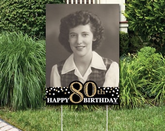 May include: Black and gold 80th birthday yard sign with a photo of a woman in a plaid shirt. The sign says "Happy 80th Birthday" with gold glitter accents.