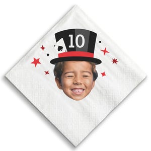 May include: White square napkin with a smiling child's face. The child is wearing a black top hat with a red band and the number 10. A playing card and red stars decorate the napkin.