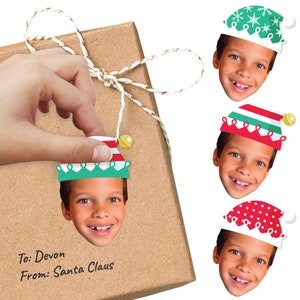 May include: Four festive Christmas gift tags featuring a child's face wearing a different elf hat. The hats are green with snowflakes, red with white stripes, green with red trim, and red with white polka dots. The text on the gift tag reads "To: Devon From: Santa Claus".