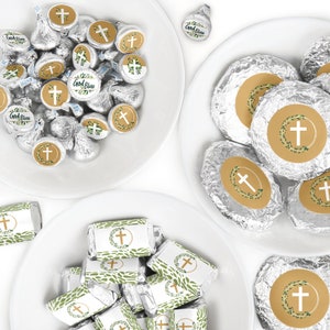 May include: An overhead shot displays white plates filled with wrapped candies. The candies feature a cross design with the words "God Bless" and are decorated with green foliage. Some candies are foil-wrapped.