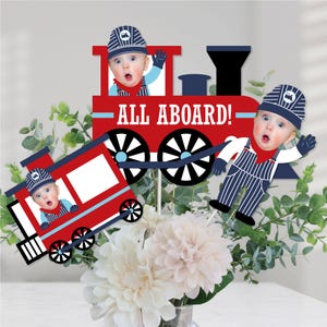 May include: Three red and blue train themed centerpieces with the text "All Aboard!" on the front of the train. Each centerpiece features a photo cutout of a baby dressed as a train conductor.