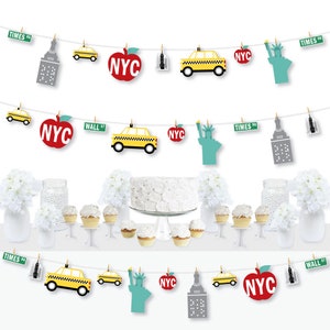 May include: A New York City themed party banner with red apples, yellow taxis, the Statue of Liberty, and the Empire State Building. The banner is strung with white twine and has small clothespins holding each piece.