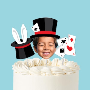 May include: A white frosted cake decorated with a smiling child's face, playing cards, and two black top hats with red bands. One hat has white bunny ears. The cake is on a light blue background.