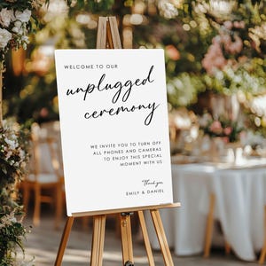 May include: A white sign on a wooden easel with the words "Welcome to our unplugged ceremony." The sign also includes a request to turn off phones and cameras. The sign is for a wedding or special event.