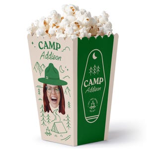 May include: A green and beige popcorn box with a camping theme. The box has a picture of a person wearing a green hat and glasses, and the text "CAMP Addison" is printed on the box.
