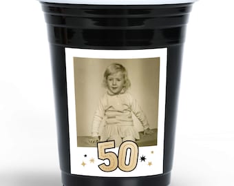 May include: A black plastic party cup featuring a rectangular white label with a sepia-toned photograph of a young child. Below the photo, the number 50 is printed in a large, beige, outlined font, surrounded by small black and gold star icons.
