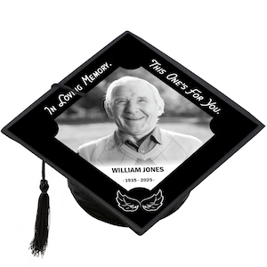 May include: Black graduation cap with a photo of a smiling older man. The cap has the text "This One's For You" and "In Loving Memory." The name "William Jones" and dates 1935-2025 are below the photo, along with a pair of angel wings.