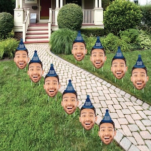 May include: A collection of eleven face cutouts with a smiling expression, each wearing a blue party hat with the number 28. The cutouts are placed in a grassy yard along a stone walkway, suggesting a birthday celebration.