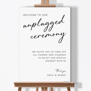 May include: A white sign on a wooden easel with black text that reads "WELCOME TO OUR unplugged ceremony." The sign also includes the text "WE INVITE YOU TO TURN OFF ALL PHONES AND CAMERAS TO ENJOY THIS SPECIAL MOMENT WITH US." and "Thank you, EMILY & DANIEL."