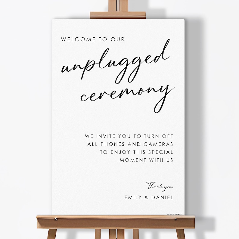 May include: A white sign on a wooden easel with black text that reads "WELCOME TO OUR unplugged ceremony." The sign also includes the text "WE INVITE YOU TO TURN OFF ALL PHONES AND CAMERAS TO ENJOY THIS SPECIAL MOMENT WITH US." and "Thank you, EMILY & DANIEL."