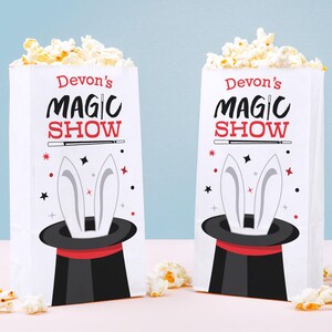 May include: Two white paper popcorn bags filled with popcorn. Each bag has the text "Devon's MAGIC SHOW" in red and black. A black top hat with white rabbit ears is featured below the text, with red and black stars.