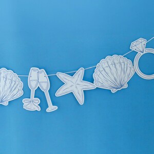 May include: A light blue and white garland featuring seashell, champagne glasses, starfish, and diamond ring cutouts. The garland is strung on a white string, perfect for a beach-themed party or wedding.