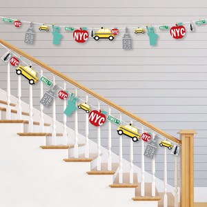 May include: A New York City themed party banner with yellow taxi cabs, red apples, and the words "NYC" and "Times Sq" hanging on a white wooden staircase.